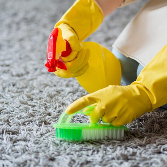 Ein grauer Teppich wird von zwei gelb behandschuhten Händen mit einer grün-weißen Bürste und einem speziellen Reiniger gereinigt.