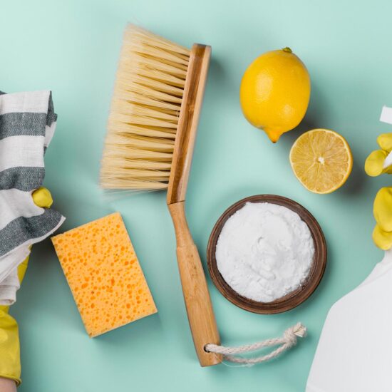 Jemand mit gelben Handschuhen hat in der linken Hand einen gestreiften Putzlappen und in der rechten Hand eine weiße Sprühflasche. Vor dieser Person liegen zwei Zitronen, eine weiße Creme, ein gelber Schwamm und ein Handfeger auf einem Türkisen Tisch